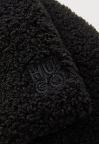 Close-up of black fleece fabric with tightly curly texture and subtle embroidered "HUGO" logo in matching black thread.