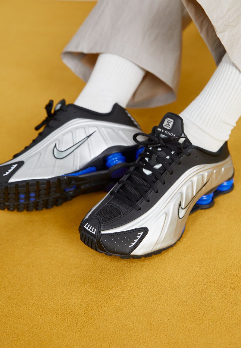 Chaussures de sport noires et argentées avec des colonnes bleu absorbant les chocs, surface texturée et lacets. Chaussettes blanches dépassant de pantalons lâches de couleur claire.