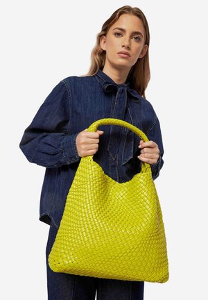 Heldergroene geweven tas in hobo-vorm met een gevlochten hengsel, gemaakt van textuurmateriaal, vastgehouden door een persoon in een spijkerblouse.