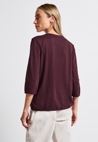 Haut bordeaux à manches trois-quarts avec un col rond et des poignets élastiqués, fabriqué dans un tissu doux et lisse, porté avec un pantalon de couleur claire.