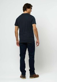 Vyras dėvi paprastą tamsiai mėlyną marškinėlį, tamsius džinsus ir rudus batus, stovintis nugara atsuktas prieš baltą foną.