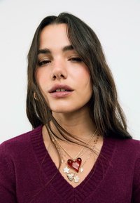 Jeune femme aux longs cheveux noirs portant des colliers en or superposés avec des charms en forme de cœur, d'étoile et de fleur, et un pull V-neck violet foncé.