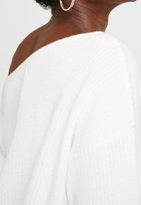 Pull blanc en maille avec un design épaule dégagée, présentant une finition texturée et une coupe ample. Comprend un accent marron à l'épaule.