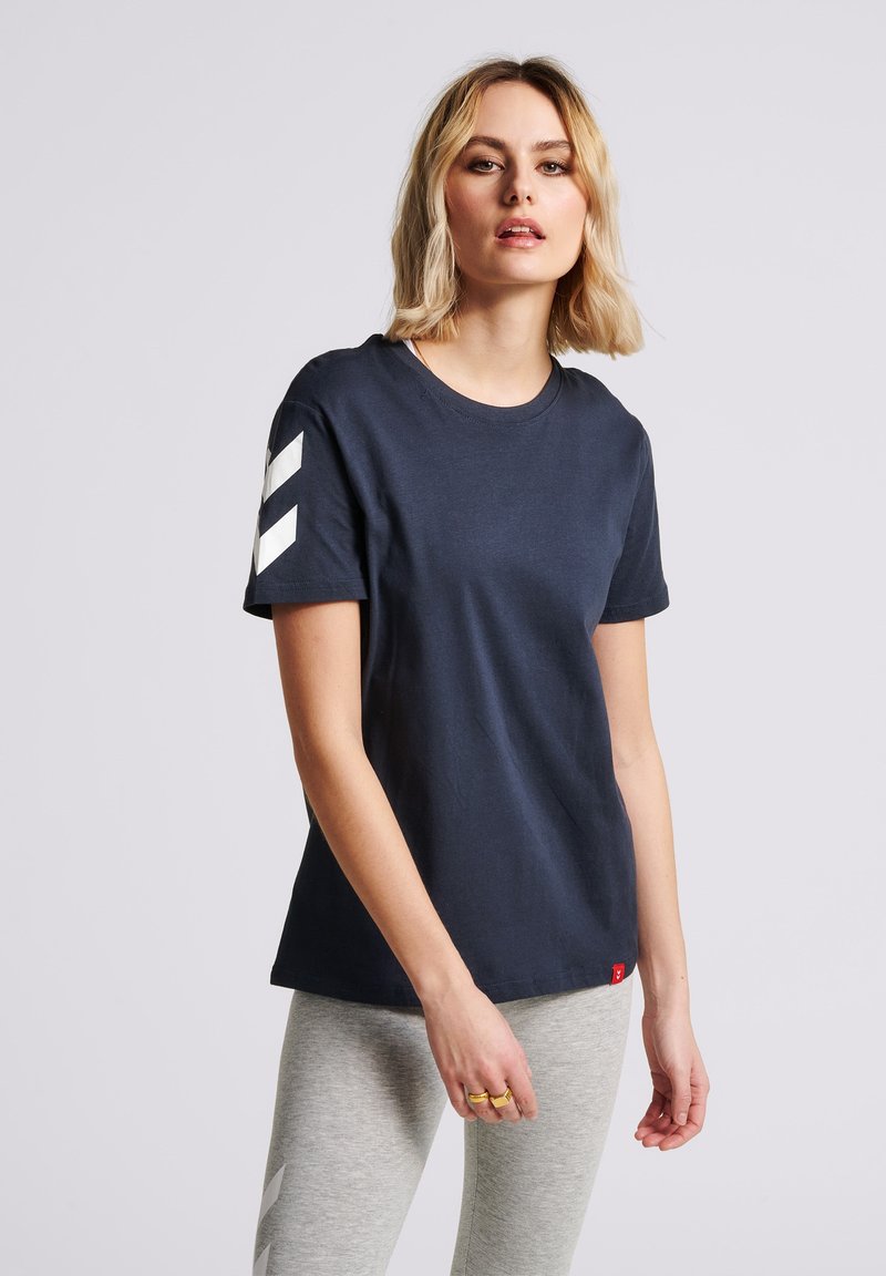 Marinblå bomulls T-shirt med rund halsringning, korta ärmar och vita chevronaccenter på vänster ärm. Har en liten röd logotag vid nederkanten.