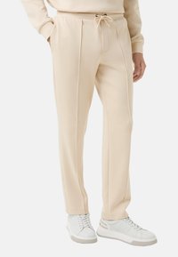 Beige schmal zulaufende Jogginghose mit einem Kordelzugbund, seitlichen Taschen und vorderen Falten, kombiniert mit weißen Sneakers. Glatte Textur.