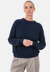 Pull en maille bleu marine avec un col rond, des poignets et un ourlet côtelés. Texture douce, coupe décontractée, associé à un pantalon de couleur claire.