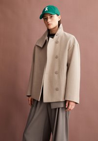 Beige wool coat with a high collar and double-breasted design. Features large buttons and relaxed fit. Paired with gray trousers and green cap.