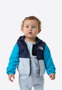 Hanorac din fleece albastru și bleumarin cu fermoar frontal, panouri contrastante și logo. Materialul interior este albastru deschis, asortat cu pantaloni albastru deschis.