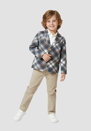 Dziecko ma na sobie szarą kratkowaną marynarkę z brązowymi i białymi akcentami, w połączeniu z białą koszulą, beżowymi spodniami i białymi trampkami.