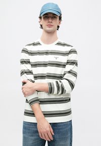 Νεαρός άνδρας με μπλε καπέλο, λευκό και μαύρο ριγέ πουκάμισο με μακριά μανίκια και μπλε τζιν, στέκεται με τα χέρια σταυρωμένα μπροστά από ένα απλό φόντο.