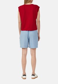 Rode katoenen mouwloze top met een ontspannen pasvorm, gecombineerd met lichtblauwe denim shorts. Model draagt witte sneakers met zwarte accenten.