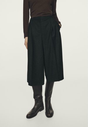 Woman wearing wide-leg black culottes, brown long-sleeve top, and knee-high black leather boots, standing with one hand in pocket.