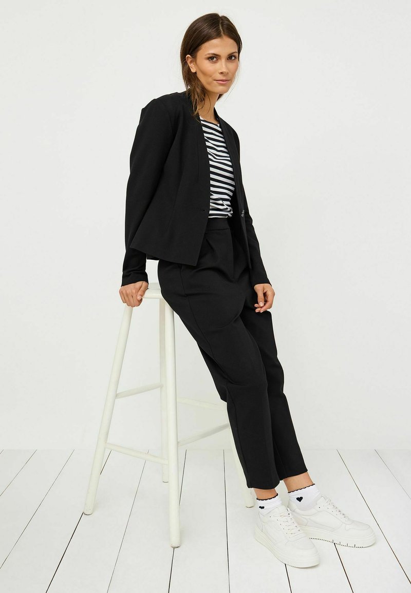 Černý přizpůsobený blejzr přes černé kalhoty a pruhované tričko, doplněný bílými teniskami a vzorovanými ponožkami, sedící na stoličce.