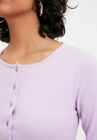 Lila pitkähihaine ylin pehmeästä kankaasta, jossa on pyöreä kaula-aukko ja viiden napin lista edessä. Sujuva rakenne ilman kuvioita.