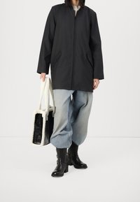 Cappotto nero fino al ginocchio con cerniera, jeans grigi over-size, stivali neri con tacco e una borsa a tracolla bianca e nera con texture.