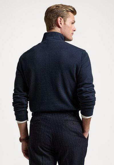 Άνδρας σε σκούρο ναυτικό πουλόβερ και ριγέ παντελόνι στέκεται με τα χέρια στις τσέπες, γυρισμένος προς τα πίσω απέναντι σε λευκό φόντο.