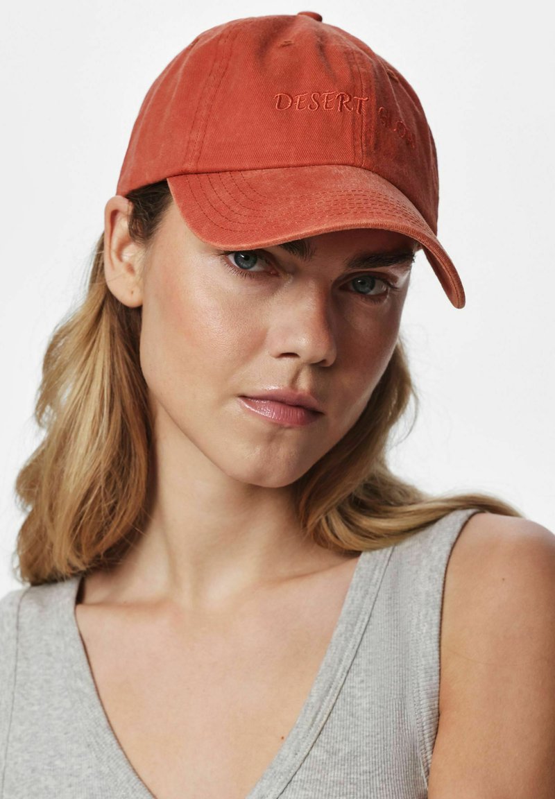 Femme à la peau claire et aux longs cheveux blonds porte une casquette de baseball rouge décolorée avec le texte "DESERT" et un haut gris sans manches sur fond blanc.