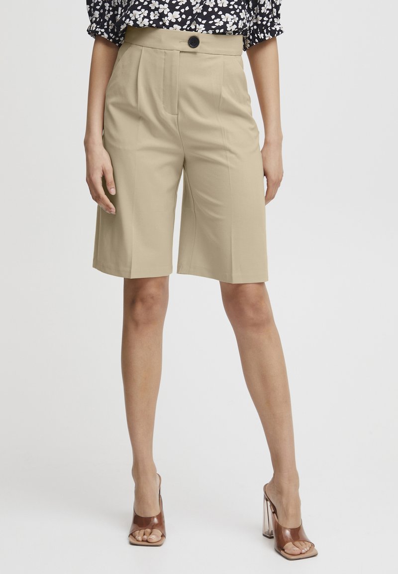 Beige maßgeschneiderte Shorts mit Vorderfalten und einem Knopfverschluss. getragen mit braunen Sandalen mit Absatz und einem schwarzen Blumenoberteil.