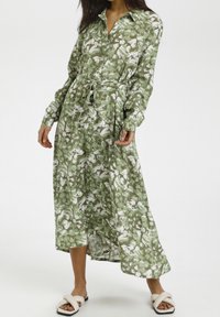 Kobieta ubrana w zieloną i białą, abstrakcyjnie wzorzystą, długą sukienkę midi z guzikami z przodu, wiązanym pasem oraz białymi, skrzyżowanymi płaskimi sandałami.