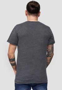 Camiseta gris de manga corta con cuello redondo, hecha de una tela suave, diseño ajustado. Tatuajes visibles en el brazo, vista trasera simple.