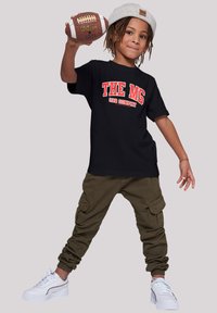 Black t-shirt with "THE MG CAR COMPANY" in red, olive cargo pants, and white sneakers with textured accents. Kid holding a football.