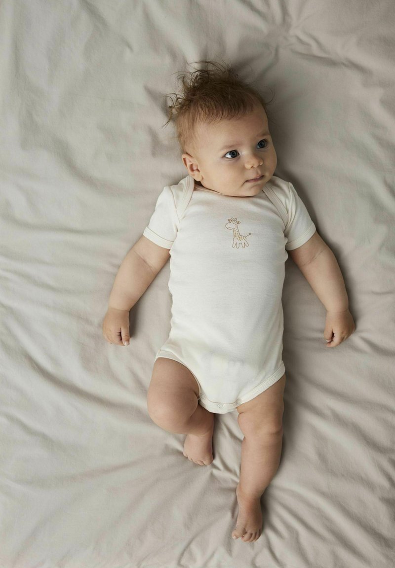 Bodysuit crème à manches courtes avec un design de giraffe, fabriqué en tissu doux, sur un couvre-lit gris clair. Bébé allongé avec une expression neutre.