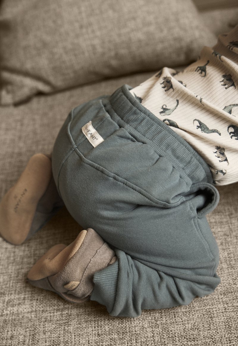 Gröna sweatpants i bomull med elastisk midjeband, texturerad sömnad och en etikett; tillsammans med beige mjuka strumpor på en texturerad yta.