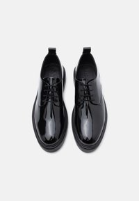 Um par de sapatos de vestir de couro preto brilhante com atacadores e solas grossas sobre um fundo cinza claro liso.