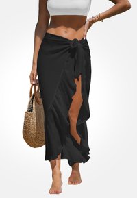 Falda maxi negra y fluida, hecha de tela ligera, con una abertura lateral y bordes con volantes. Nudo atado en la cintura. Bolso de rafia en la mano.