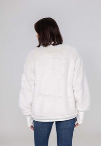 Witte nepbontjas met een ontspannen pasvorm, ribgebreide manchetten en een zachte textuur, met een ronde halslijn en een elastische zoom. Draagt over blauwe jeans.