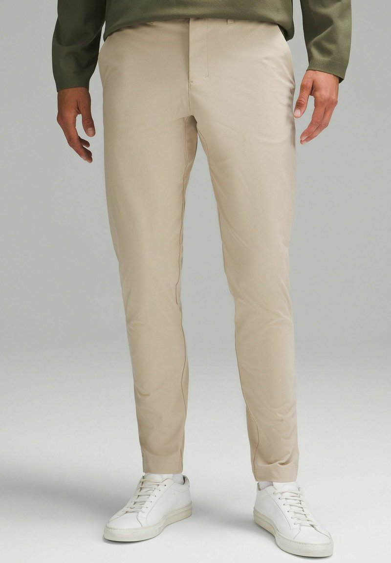 Beżowe spodnie slim-fit wykonane z gładkiego materiału, z płaskim przodem i kieszeniami po bokach, połączone z białymi trampkami.