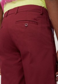 Burgundy byxor gjorda av vävt tyg, med en bakficka med knappstängning och subtil logobroderi. Slät textur.