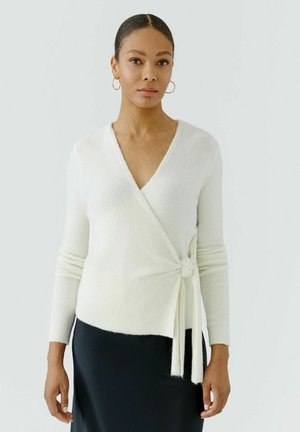 Frau mit zurückgebundenem Haar, die goldene Creolen, einen weißen Wickelpullover, der an der Taille gebunden ist, und einen schwarzen Rock trägt, steht vor einem hellen Hintergrund.