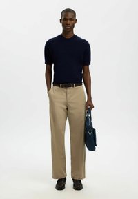 Chemise en maille bleu marine à manches courtes, pantalon large kaki, ceinture tressée marron et chaussures texturées noires, tenant un sac bleu avec un accent triangulaire.