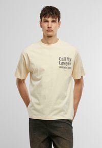 Lys gul bomulds t-shirt med rund hals, med et trykt grafik i mørkegrå på forsiden, inklusive tekst og telefonnummer.