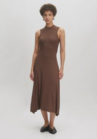 Braunes geripptes ärmelloses Kleid mit tailliertem Oberteil und schwingendem Rock, ausgestattet mit einem Rundhalsausschnitt und asymmetrischem Saum. Schwarze Schuhe.
