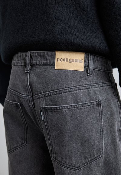 Jeans gri din denim cu un patch din piele etichetat „noongoons”. Designul include cinci buzunare și detalii de cusătură vizibile.