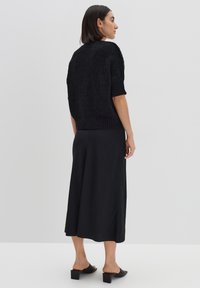 Schwarzer gestrickter Pullover mit kurzen Ärmeln, kombiniert mit einem langen schwarzen Rock. Das Outfit zeichnet sich durch eine lockere Passform und eine weiche Textur aus. Schwarze Schuhe mit Blockabsatz.