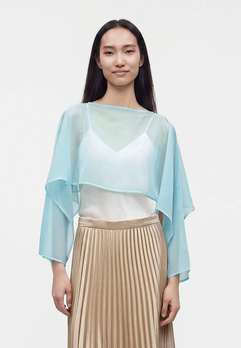 Femme aux longs cheveux foncés porte un top bleu clair transparent sur un caraco blanc et une jupe beige plissée, debout devant un fond uni.