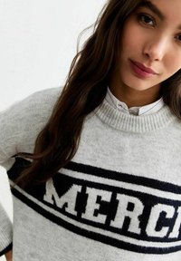 Grå stickad tröja med ett svart och vitt randigt mönster och "MERCI"-text över framsidan. Ribbad halsringning och mudd.