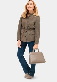 Braune Jacke aus weichem Stoff mit einem taillierten Bund, zwei vorderen Taschen und Knopfverschluss; kombiniert mit blauen Jeans und einer grauen Handtasche.