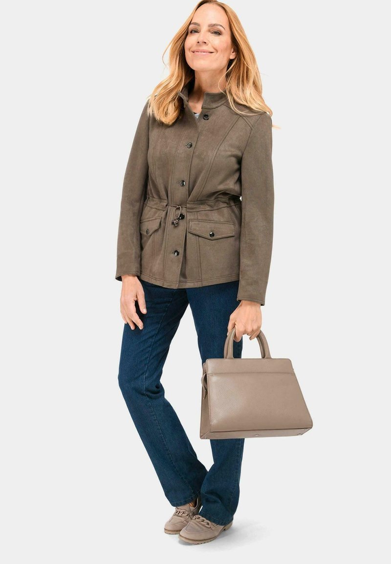 Braune Jacke aus weichem Stoff mit einem taillierten Bund, zwei vorderen Taschen und Knopfverschluss; kombiniert mit blauen Jeans und einer grauen Handtasche.