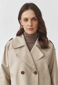Beež trench coat topeltroolide nuppudega, suurte kraede ja õlavarre epauletidega. Kandy pruunist soonikust turtlenecki küljes. Sile tekstuur.