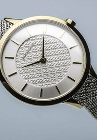 Round silver watch with a patterned dial, gold accents, and a mesh strap. Features black hour and minute hands. Clean, modern design.