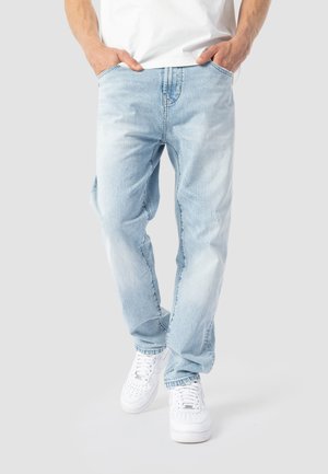 Mann trägt hellblaue Jeans und weiße Sneaker mit den Händen in den Taschen vor einem einfarbigen Hintergrund.