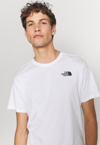 Camiseta blanca de algodón con mangas cortas, que presenta un pequeño logo negro en el pecho. Textura suave, cuello redondo clásico. Diseño sencillo.