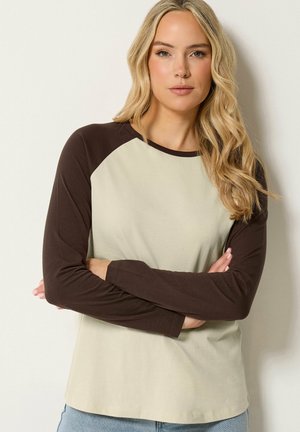 Mujer rubia con cabello ondulado, que lleva una camisa beige de manga larga con mangas marrones oscuras, de pie con los brazos cruzados frente a un fondo claro.