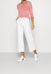 Rosa Langarmtop mit einem bestickten Ausschnitt, weißen, weit geschnittenen, verkürzten Hosen und weißen Sneakers mit schwarzen Akzenten. Glatte Textur und lässige Passform.