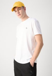 Camiseta de algodón blanca con un pequeño logo en el pecho, combinada con pantalones negros. El modelo lleva una gorra amarilla brillante.