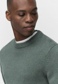 Vihreä neulepaita pyöreällä pääntiellä ja ribbineulehuuliilla. Pehmeä rakenne hienovaraisilla yksityiskohdilla, sijoitettuna vaaleaa taustaa vasten.